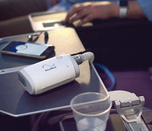 A portable ResMed CPAP machine on an airplane tray table next to a smartphone. A passenger's hand is visible in the background